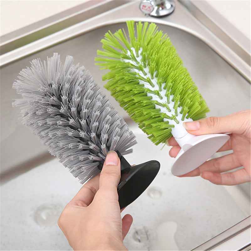 Two bottle brush cleaning tools being held over a stainless steel sink. One brush has gray bristles and a black base, and the other has bright green bristles with a white center and a white suction-style handle.