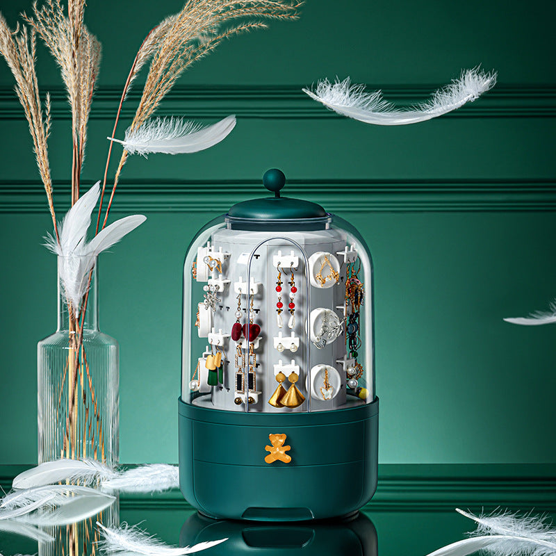 A dark teal jewelry display stand with a clear dome top, filled with earrings and jewelry pieces, set against a dark green paneled background with decorative white feathers and a glass vase of dried grass.