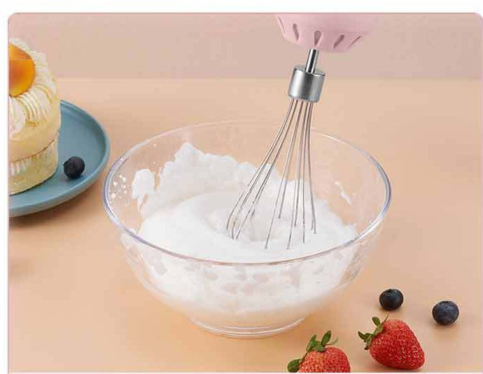 A pink electric hand mixer whisk attachment shown mixing white cream in a clear glass bowl, with a small cake on a blue plate and strawberries and blueberries nearby on a peach-colored surface.