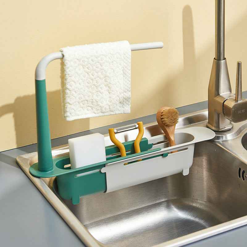 Kitchen sink caddy with green holder, white sponge, yellow scrubber, and brush on a stainless steel sink.