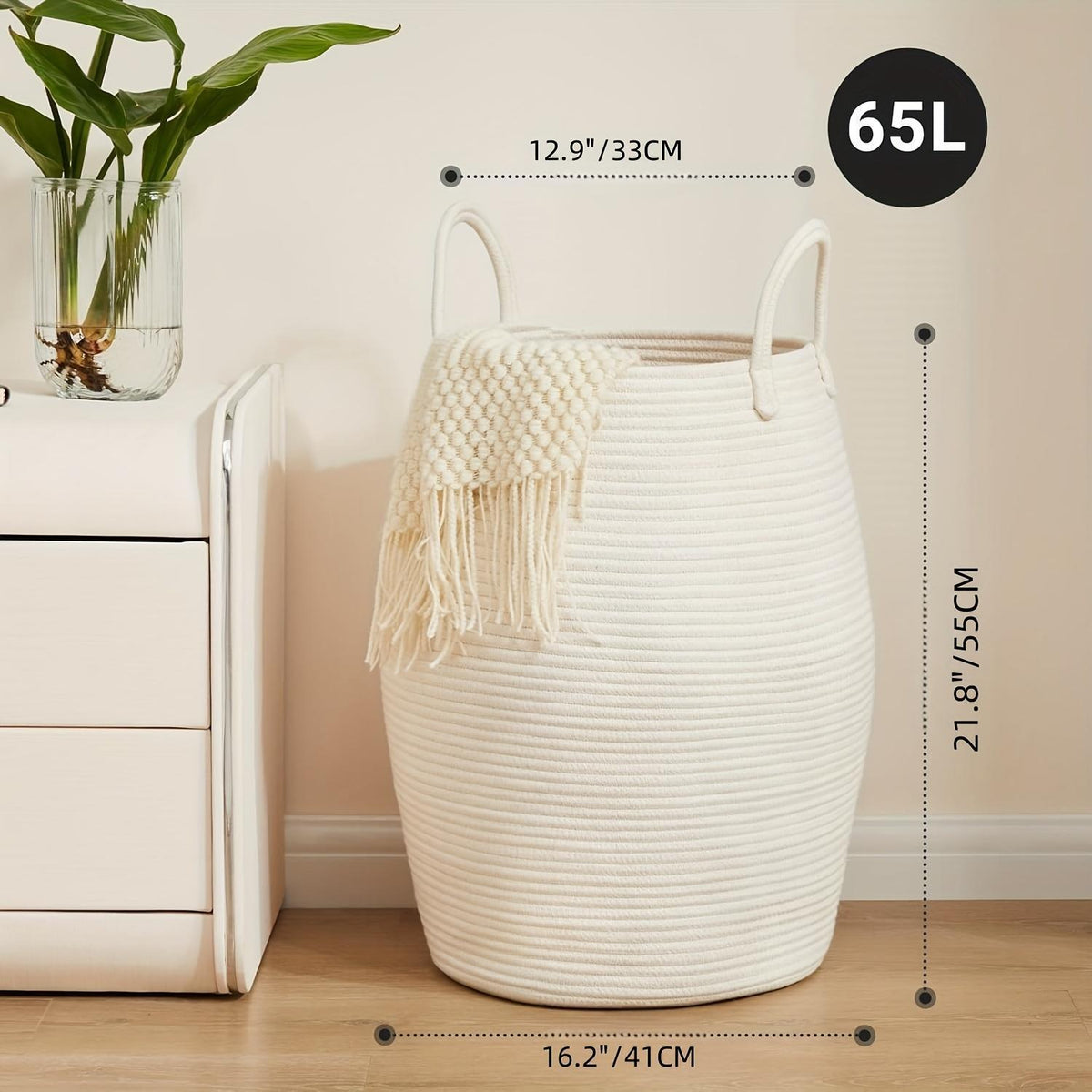 White woven basket with handles on a wooden floor, next to a white cabinet and plant, with measurements displayed.