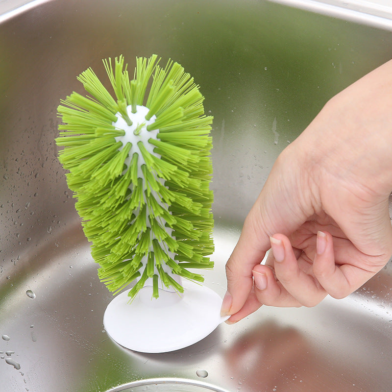 A green bottle brush with a white circular base is shown inside a stainless steel sink, with a hand holding the base.