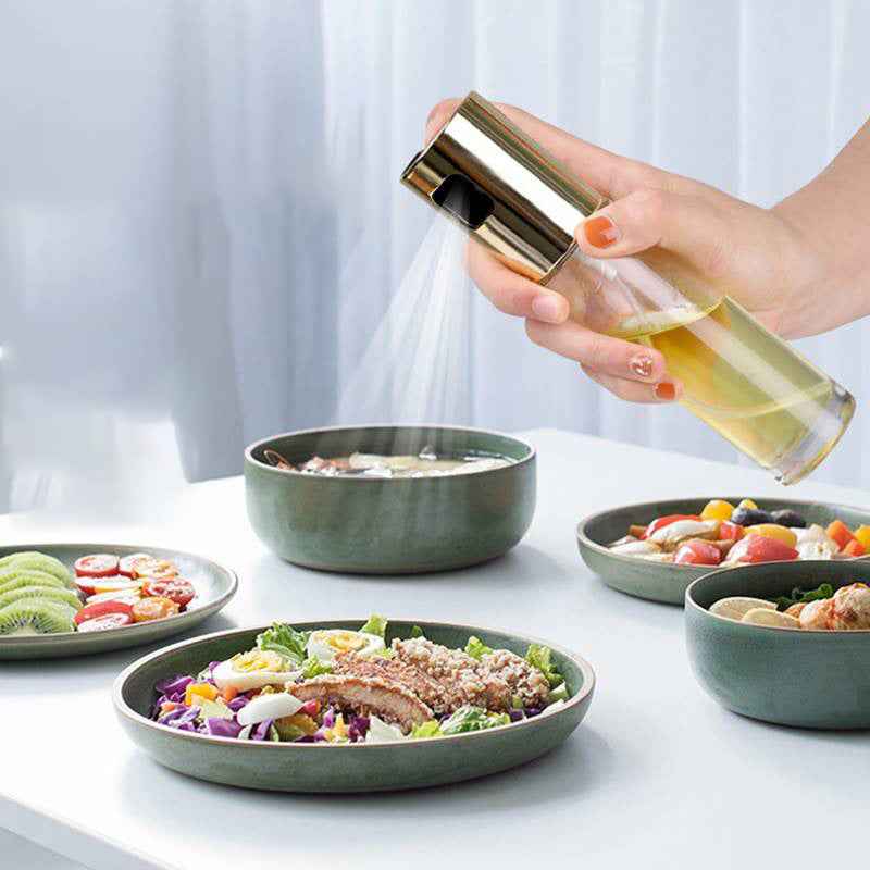 A hand holding a clear bottle with a shiny gold sprayer dispenses oil over a salad in a green plate, with other prepared dishes on a table in a bright indoor setting.