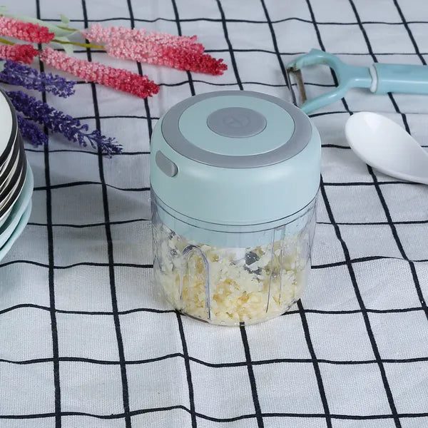 A small electric food chopper with a light blue lid and clear container holding chopped garlic, placed on a black-and-white checkered cloth with kitchen props like a peeler, spoon, and flowers.