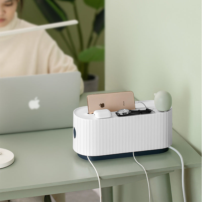 A white ribbed cable organizer desk stand holding a smartphone, earbuds case, and small accessories, placed on a light green desk in a home office setting with a blurred person in the background.