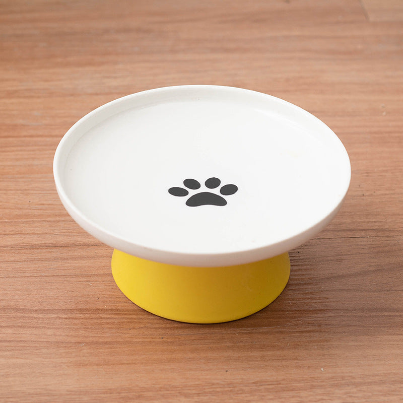 A white ceramic pet food bowl with a black paw print graphic in the center, resting on a yellow silicone base over a wooden tabletop.