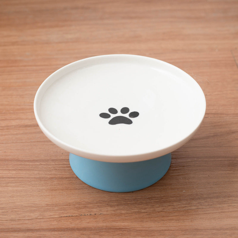 A pet elevated food bowl with a white ceramic plate and a light blue silicone base, featuring a black paw print graphic in the center, set on a wooden surface.