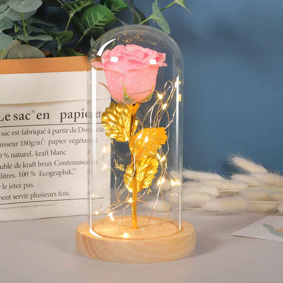 Pink and gold rose in a glass dome with LED lights on a wooden base, placed on a surface with a blurred background.