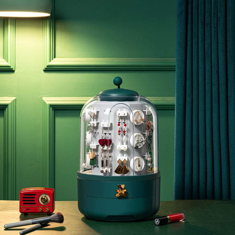 A teal jewelry display cabinet with a clear dome showing earrings and jewelry inside, placed on a tabletop with a red vintage radio, makeup brushes, and lipstick nearby against a green paneled wall and dark blue curtain.
