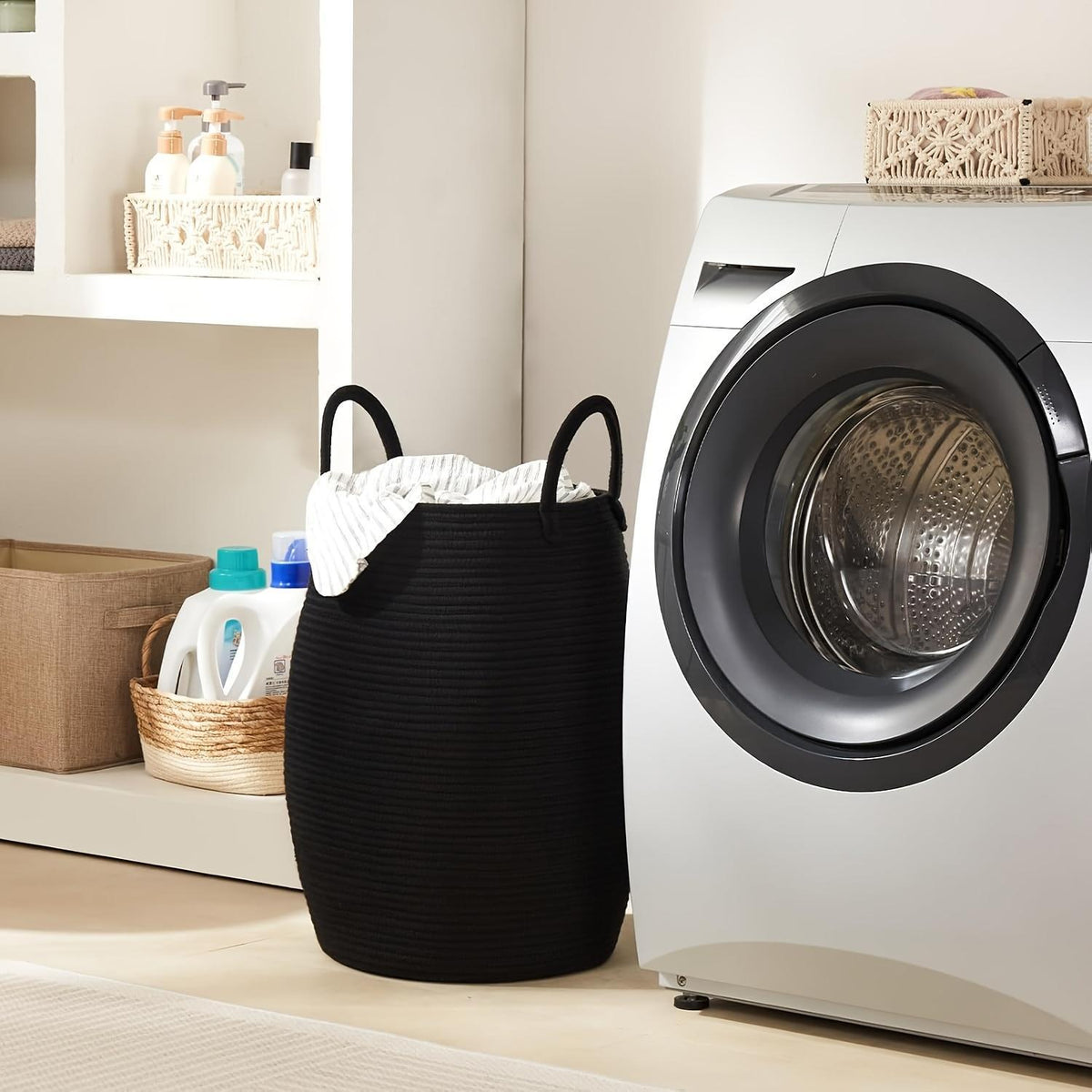 A black ribbed fabric laundry hamper with two handles, filled with white clothes, standing next to a white front-loading washing machine in a bright laundry room.