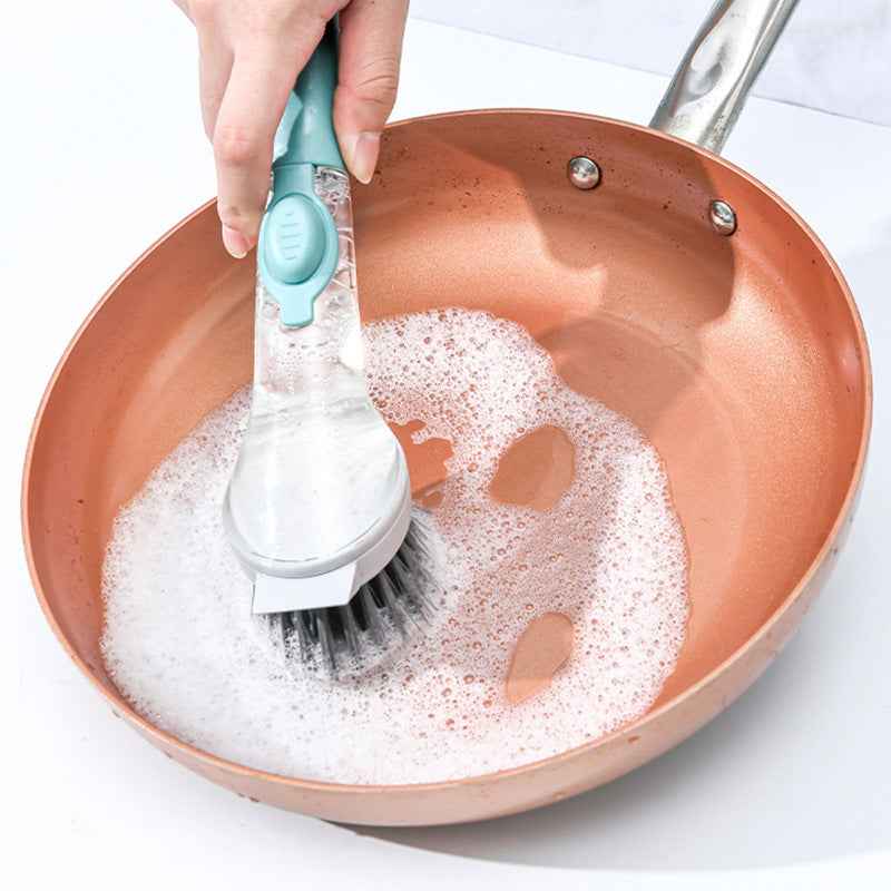 A hand holds a silicone cleaning brush with black bristles and a mint green handle, scrubbing a copper-colored pan filled with soapy foam on a white surface.