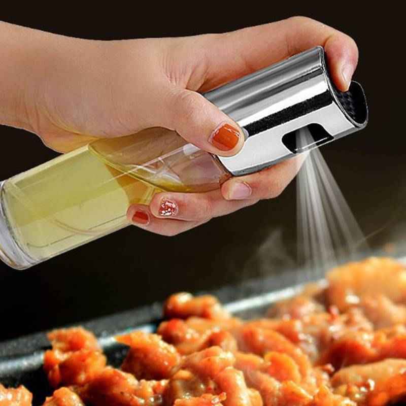 Hand holding a bottle of oil and a metallic spray nozzle over food on a grill.