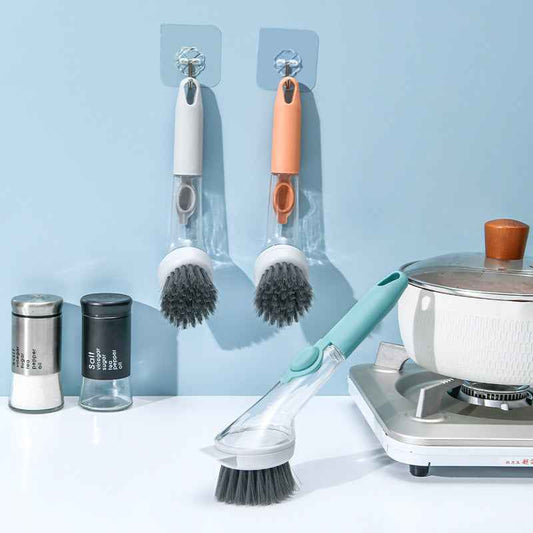 Two wall-mounted kitchen cleaning brushes with clear handles and dark bristles are shown hanging on a light blue wall above a countertop. A second brush with a mint green handle rests on the counter, alongside a pot on a small burner and two salt and pepper shakers.