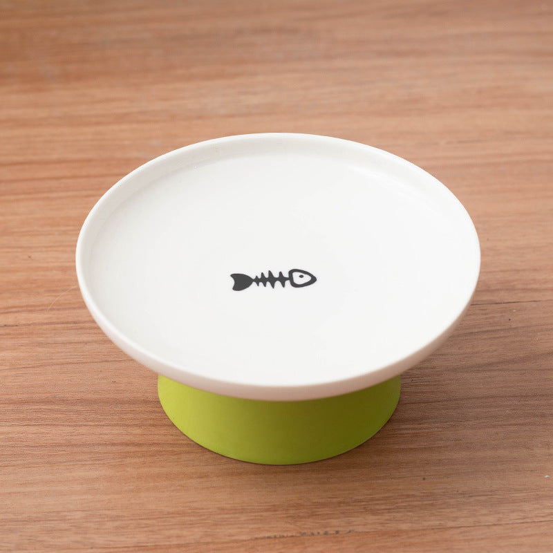 A white ceramic pet bowl with a black fish skeleton graphic in the center, resting on a light green silicone base over a wooden tabletop.