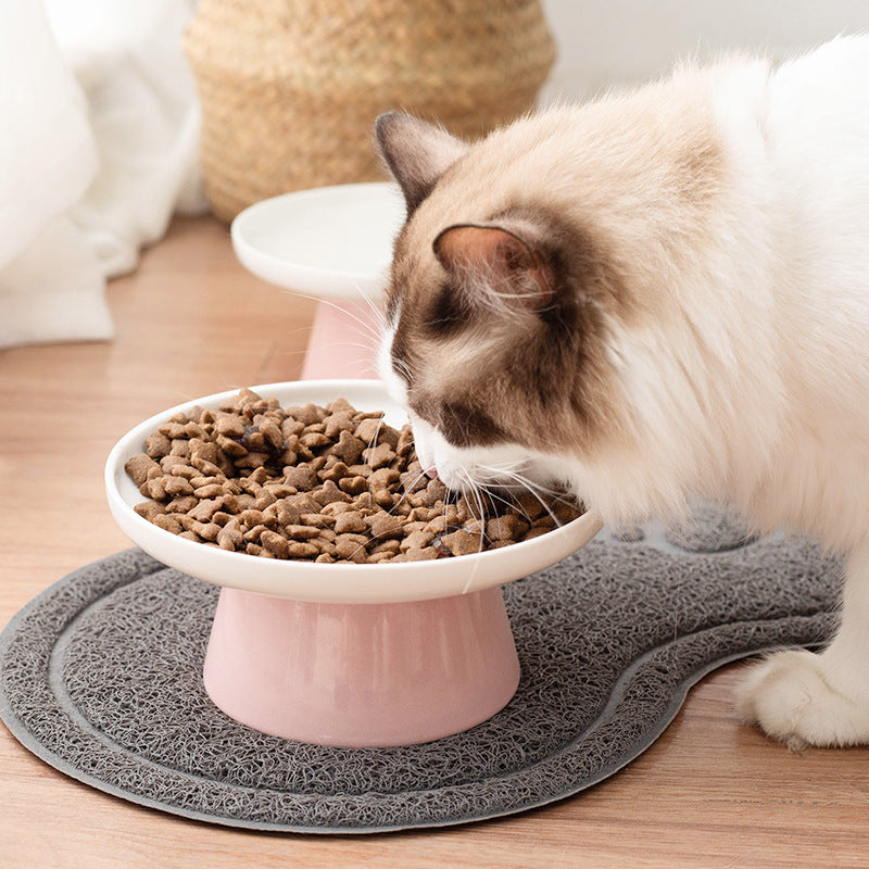 A cat with cream, white, and brown fur is eating dry kibble from a pink and white elevated pet bowl placed on a gray textured mat on a wooden floor.