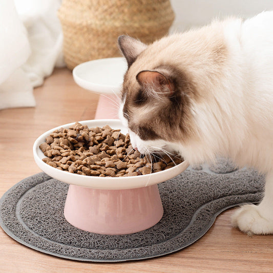 A cat with cream, white, and brown fur is eating dry kibble from a pink and white elevated pet bowl placed on a gray textured mat on a wooden floor.