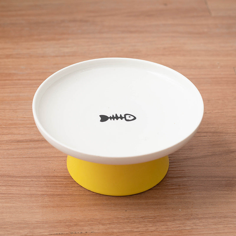 A white elevated pet food bowl with a yellow base, featuring a simple black fish skeleton icon printed in the center of the bowl, set on a wooden surface.
