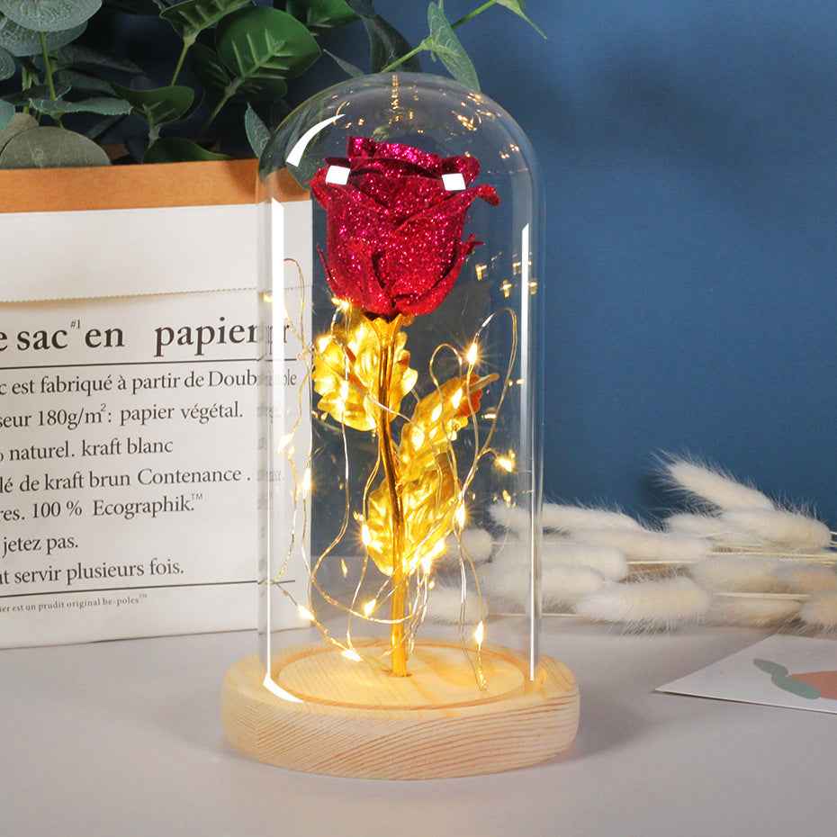 Red rose with gold base and string lights inside a glass dome on a wooden base.