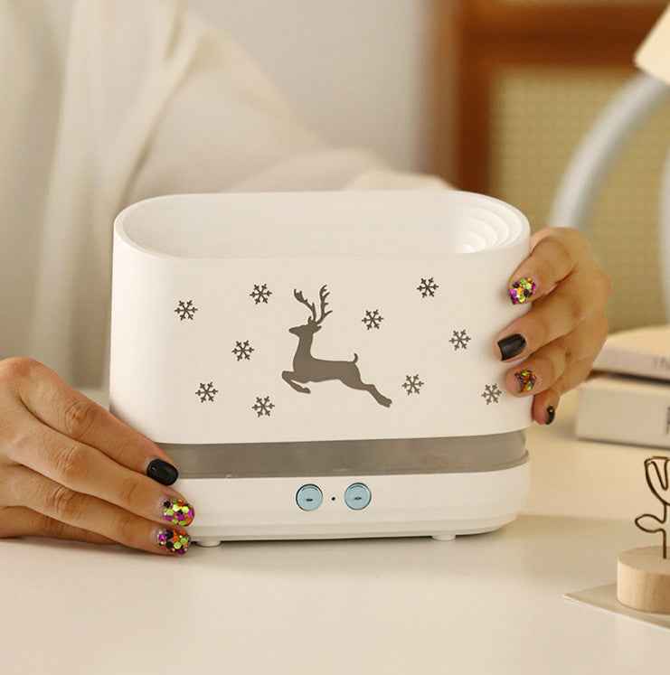 A white rectangular humidifier or diffuser with a gray deer and snowflake design, held by hands with colorful nail art on a light-colored table.