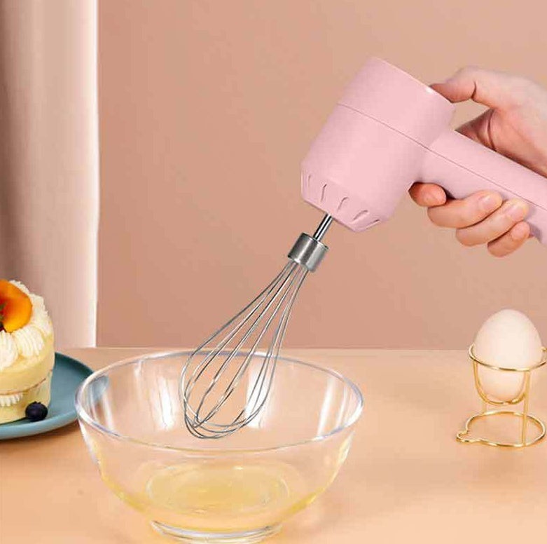 A pink handheld electric hand mixer with a whisk attachment being used to beat an egg in a clear glass bowl, set against a soft peach background with a small cake and egg holder in the background.
