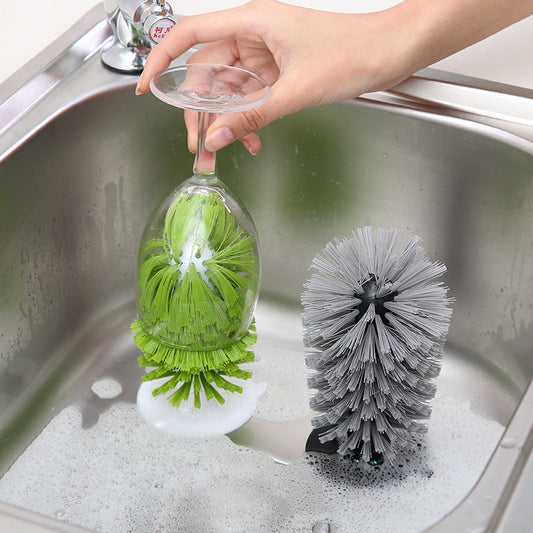 A hand holds a clear wine glass over a stainless steel sink while a green cleaning brush sits inside the glass. Another gray cleaning brush is placed in the sink, which contains soapy water.