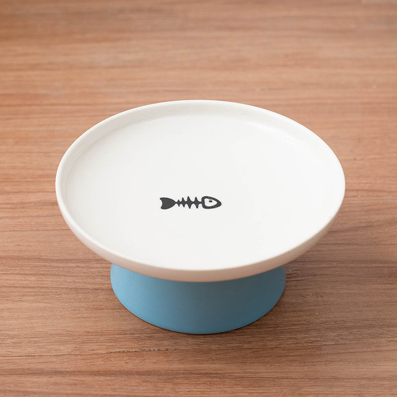 A white ceramic pet food bowl with a blue silicone base, featuring a black fish skeleton icon printed on the inside of the bowl, set on a wooden tabletop.
