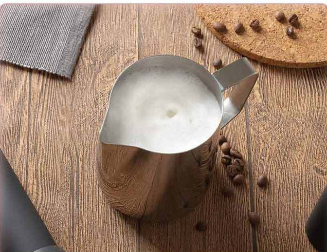 Metal pitcher pouring milk into a glass on a wooden surface with coffee beans.