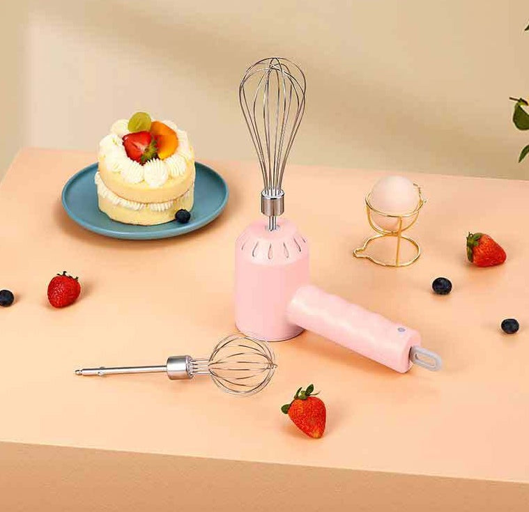 A pink handheld electric hand mixer with two metal whisk attachments, styled on a peach tabletop with a decorated cake, strawberries, blueberries, and a gold egg stand in the background.