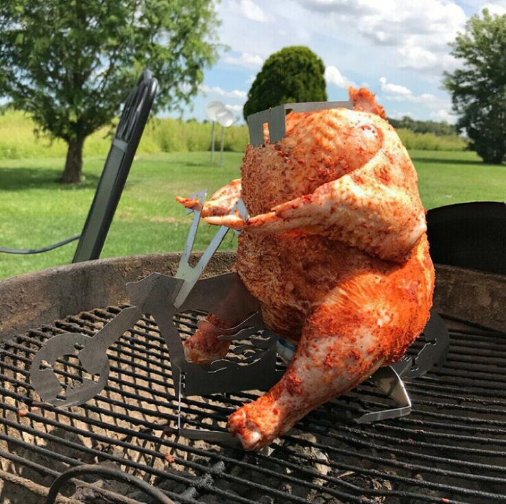 A whole raw chicken coated in orange-red seasoning, mounted on a metal rotisserie stand on a grill grate outdoors with a grassy field and trees in the background.