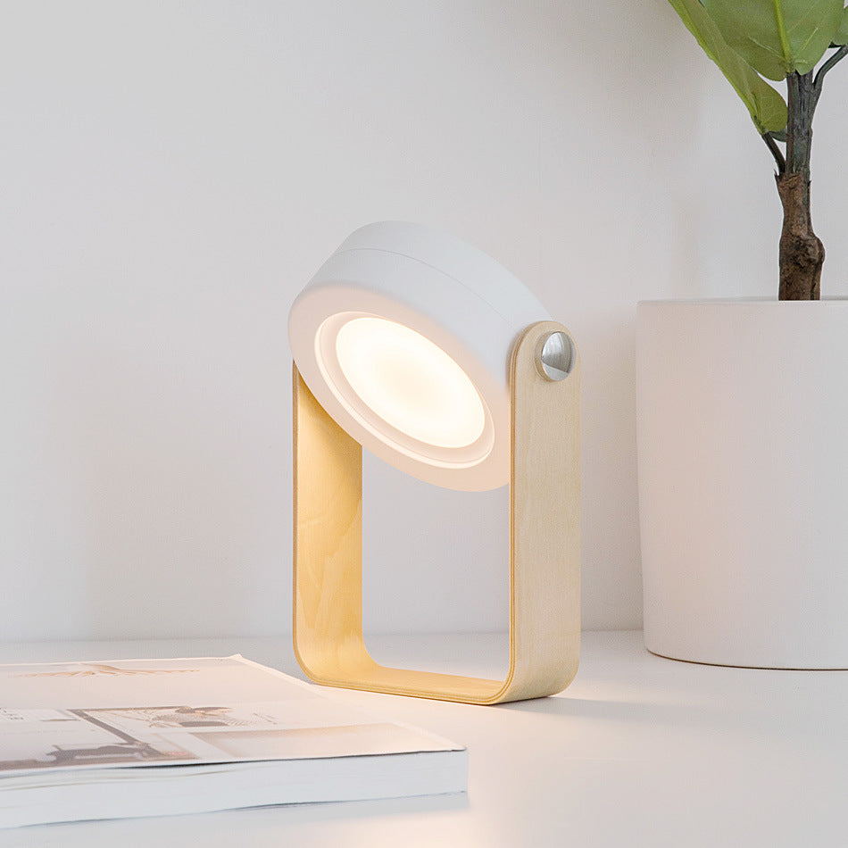 Modern table lamp with wooden frame and white base on a surface with a plant in the background.
