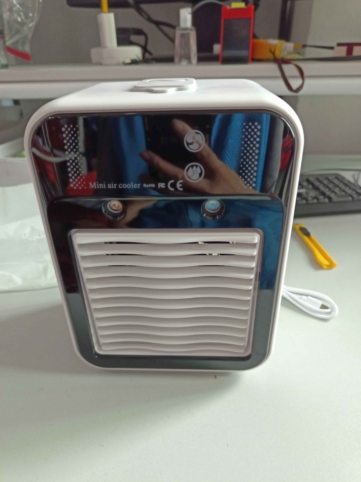 A small portable mini air cooler with a white plastic body, a glossy black front panel, and a white vented grille on the front. It is shown sitting on a white desk with its power cord coiled next to it, with a blurry office background.