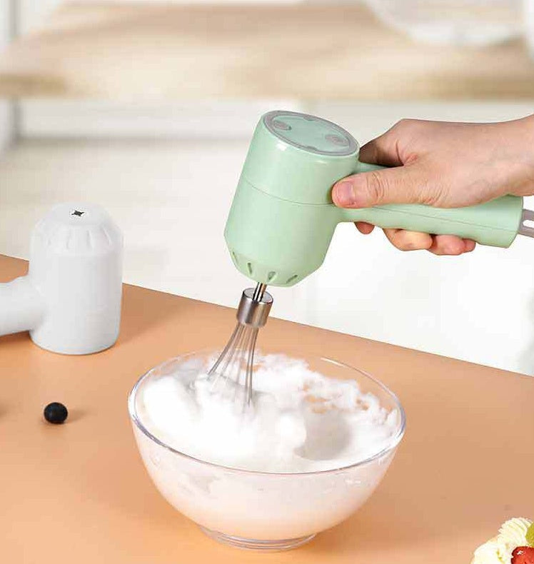 A hand holding a mint green handheld electric mixer with a whisk attachment, beating white foam in a clear glass bowl on a peach-colored table. A white container is visible in the background.