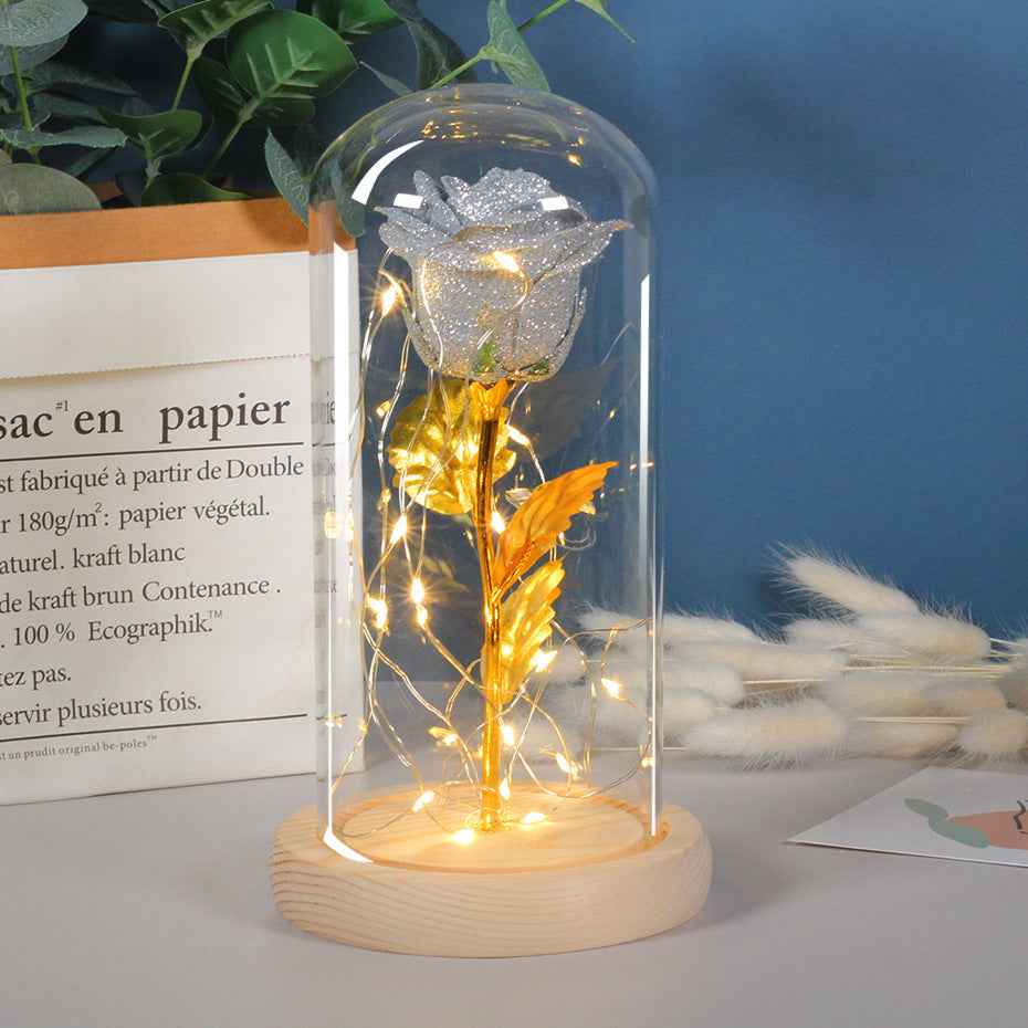 Decorative rose in a glass dome with LED lights on a wooden base, placed on a surface with a book and plants in the background.
